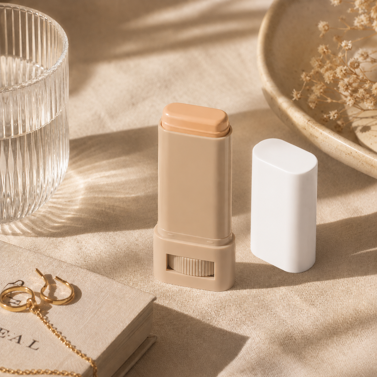 Beige makeup stick with white cap on a beige surface with a glass and dish in the background